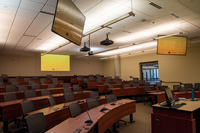 View from front left of classroom showing confidence panels, audience seating and mics, room camera, and projectors.  Front podium and monitor is visible on the right.