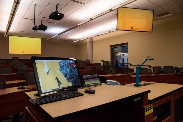 View from behind instructor podium.  Monitor and A/V control panel are visible along with rear project, confidence panel, audience seating and mics, and document camera.  
