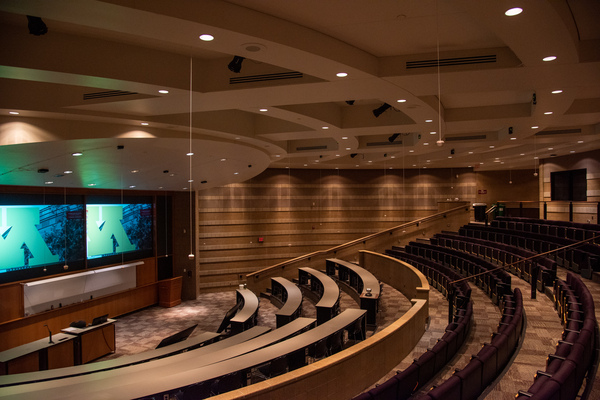 Audience view from rear left.  Most of the audience seating spread out in auditorium style is visible, along with student mics hanging from the ceiling. Front projector displays, front white board, and emergency exit are also visible.